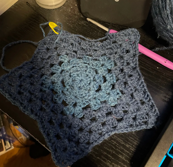 a blue star coaster in the granny square stitch on my desk, it has a yellow stitch marker in it and a pink crochet hook attached to the side